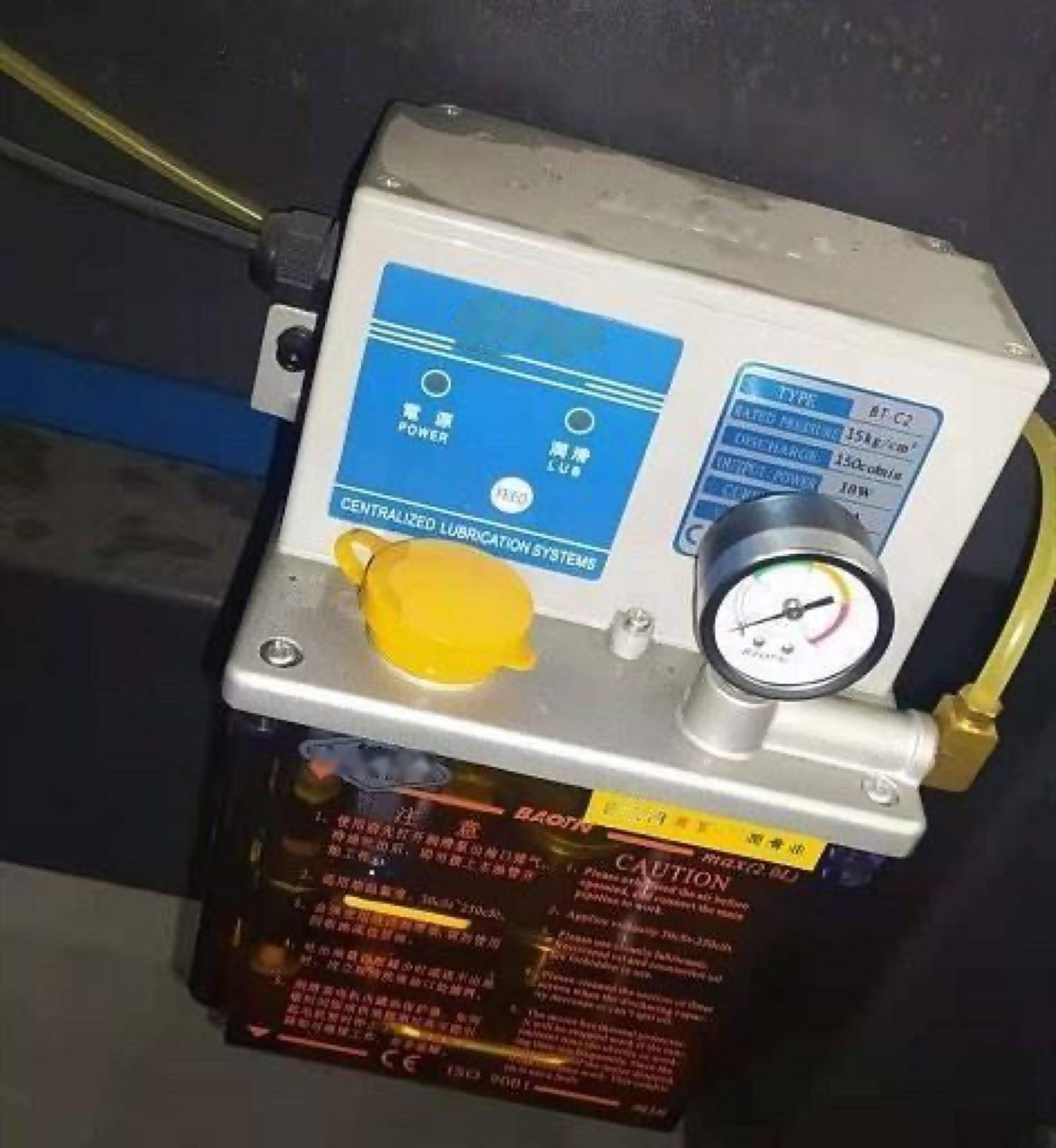 Centralized lubrication pump with an oil tank, a pressure gauge and a yellow filler cap, mounted on the machine